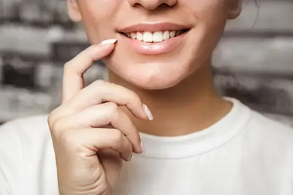 A close-up of a woman’s smile as she gently touches her teeth with her finger, highlighting white and healthy teeth.