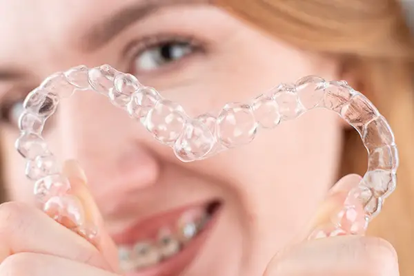 Smiling woman with braces holding clear aligners in a heart shape, showcasing orthodontic solutions.