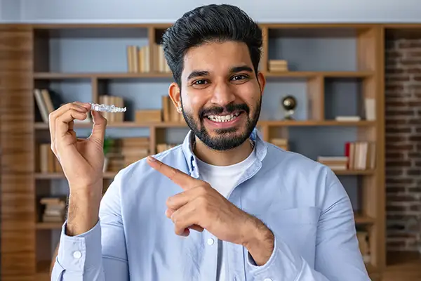 Smiling man holding and pointing at a clear aligner in a home setting, highlighting its use for adult teeth alignment.