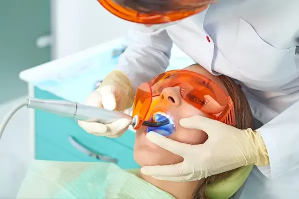 A dentist using a curing light to set a dental filling while the patient wears protective orange glasses during the procedure.