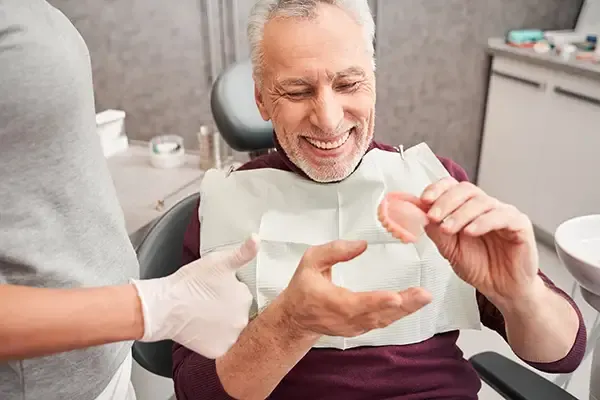 Elderly male patient smiling and examining a set of dentures during a consultation at a dental clinic.