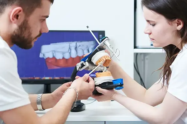 Two dental technicians using a model to design and prepare for full-mouth dental restoration, with 3D imaging on a monitor in the background.