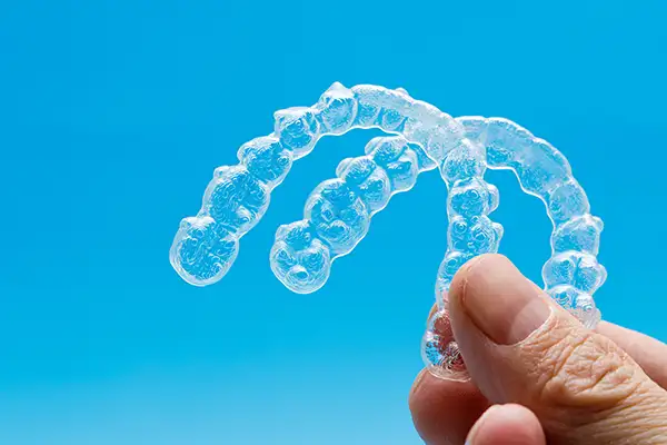 Close-up of a hand holding upper and lower Invisalign clear aligner trays, the blue background shining through to showcase the trays’ transparent, light material.